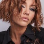 Model wearing a short, wavy, auburn wig with side-swept bangs.